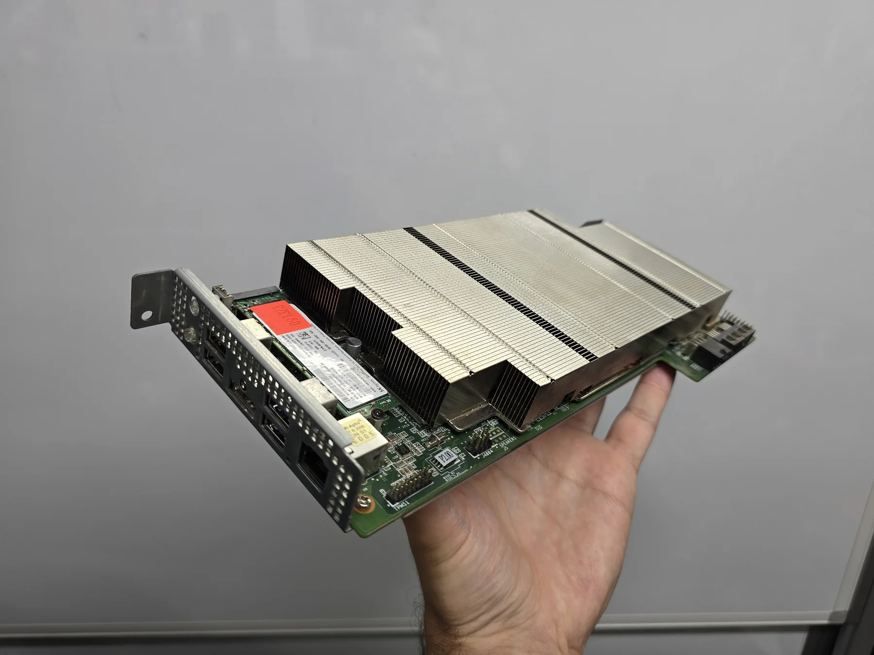 An actual BC-250 held in one hand, showing its silver heatsink, installed SSD and rear ports